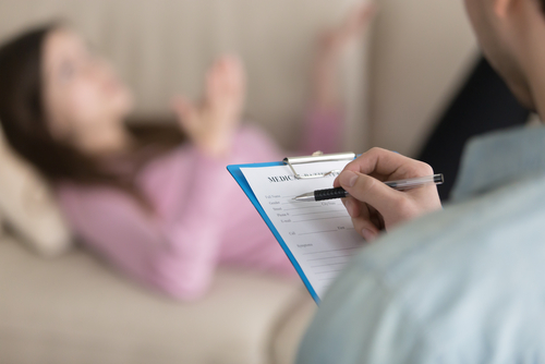 male psychotherapist holding clipboard, filling information about patient while lady lying on couch in the background