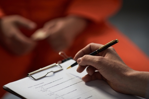 Caucasian woman holding pen and clipboard conducting interview with prisoned adult wearing orange uniform