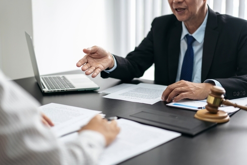 A man and a woman are sitting at a table with papers and a laptop. The man is wearing a suit and tie